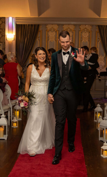 A happy couple walks down the aisle as guests applaud in a warmly lit hotel ceremony.