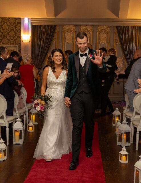 A happy couple walks down the aisle as guests applaud in a warmly lit hotel ceremony.