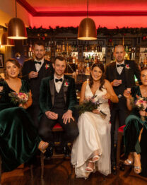 A group of well-dressed adults, some holding drinks, gather in a warmly lit hotel bar adorned with festive decor.
