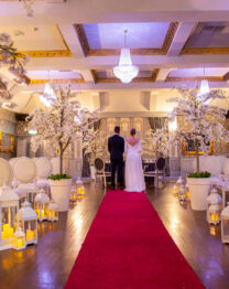 A couple stands in an elegantly decorated hotel hall with white blossoms and a red carpet.