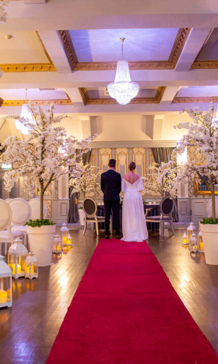 A couple stands in an elegantly decorated hotel hall with white blossoms and a red carpet.
