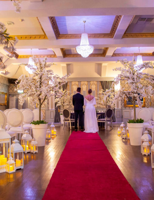 A couple stands in an elegantly decorated hotel hall with white blossoms and a red carpet.