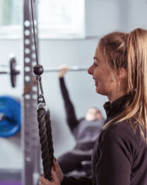 A woman exercises in a bright gym, with a man lifting weights in the background.