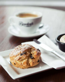 Close-up of a scone with cream, with two coffees on a wooden table.