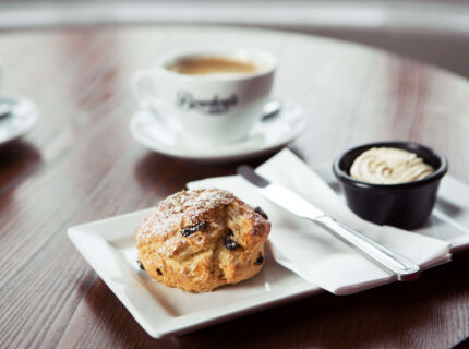 Close-up of a scone with cream, with two coffees on a wooden table.