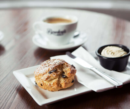 Close-up of a scone with cream, with two coffees on a wooden table.