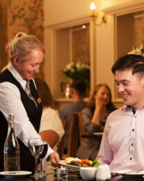 Friendly waitress serves dinner to smiling guest in warm, elegant hotel restaurant setting.
