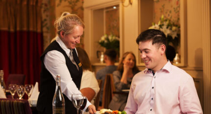 Friendly waitress serves dinner to smiling guest in warm, elegant hotel restaurant setting.