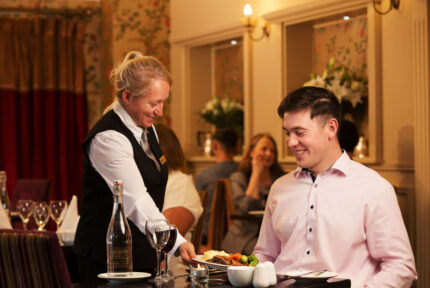 Friendly waitress serves dinner to smiling guest in warm, elegant hotel restaurant setting.