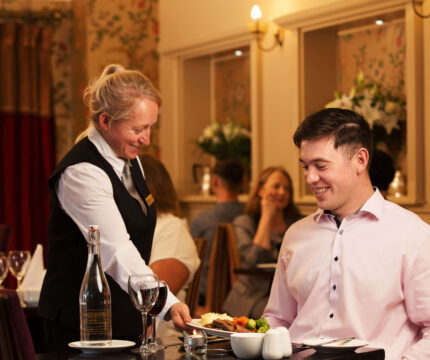 Friendly waitress serves dinner to smiling guest in warm, elegant hotel restaurant setting.