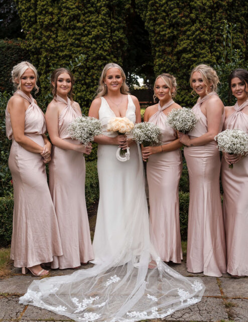 A joyful bridal party in a garden at The Anner Hotel, wearing soft pink dresses.
