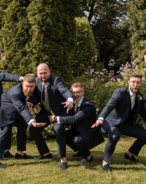 A joyful group of men in suits posing playfully outdoors against lush greenery.