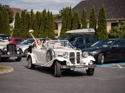 A joyful couple drives a classic wedding car outside The Anner Hotel's lush conference centre.