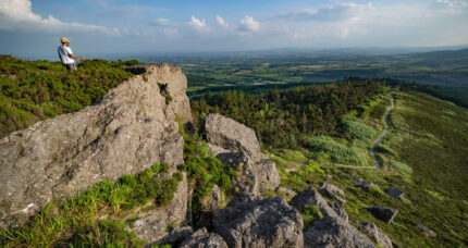 Lone hiker enjoys stunning view from rocky hilltop, with vast lush landscape and clear blue sky.
