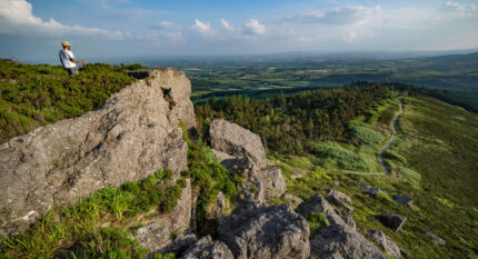 Lone hiker enjoys stunning view from rocky hilltop, with vast lush landscape and clear blue sky.