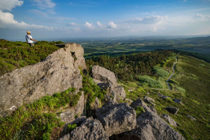 Lone hiker enjoys stunning view from rocky hilltop, with vast lush landscape and clear blue sky.