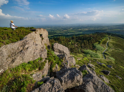 Lone hiker enjoys stunning view from rocky hilltop, with vast lush landscape and clear blue sky.