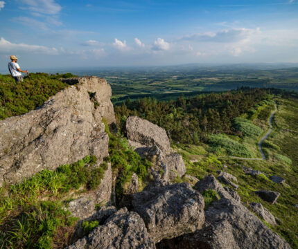 Lone hiker enjoys stunning view from rocky hilltop, with vast lush landscape and clear blue sky.
