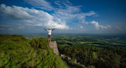 A person stands on a cliff edge enjoying a vast, scenic countryside view under a blue sky.