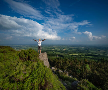 A person stands on a cliff edge enjoying a vast, scenic countryside view under a blue sky.