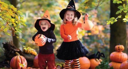 Two children in Halloween costumes holding pumpkins in an autumnal garden setting.
