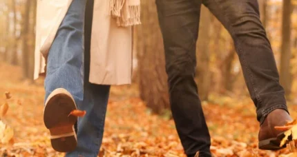 A couple strolls hand-in-hand through an autumnal, leaf-strewn woodland.