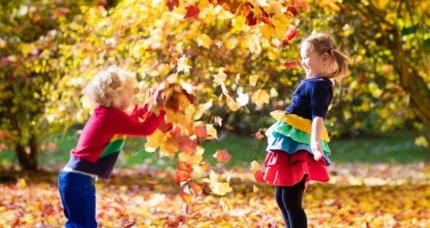 Two children joyfully play in autumn leaves at a park, surrounded by vibrant fall foliage.